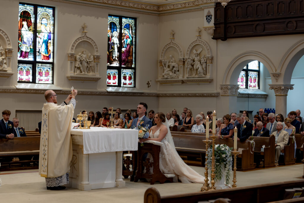 Priest elevates the Eucharist as the Catholic couple and congregation kneels in adoration.
